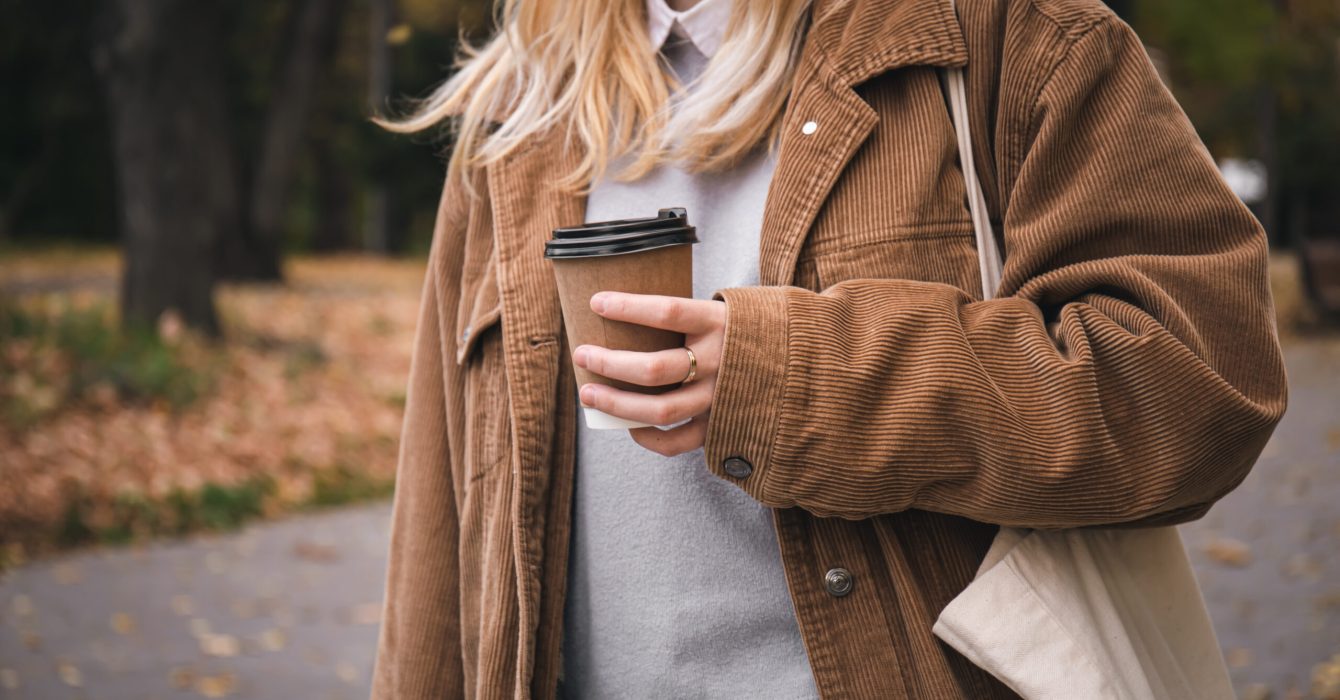 A young woman holds a craft disposable glass of coffee in her hand on a walk in the park, close up.
